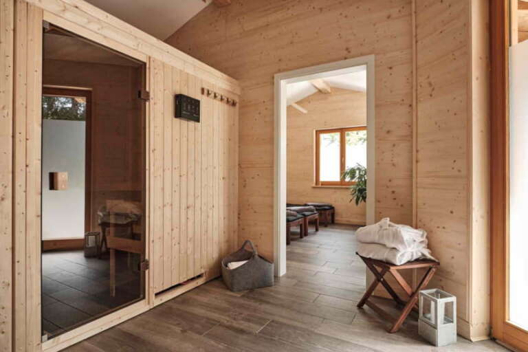 Die moderne Holzsauna mit Glastür im Hotel Hochfirst verfügt über einen Handtuchhalter, einen Korb und einen durch eine offene Tür einsehbaren Ruhebereich mit Liegestühlen.