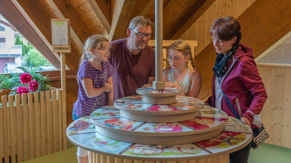 Familie mit zwei Töchtern betrachtet gemeinsam eine runde interaktive Bilderscheibe in einer Ausstellung mit Holzdachkonstruktion.