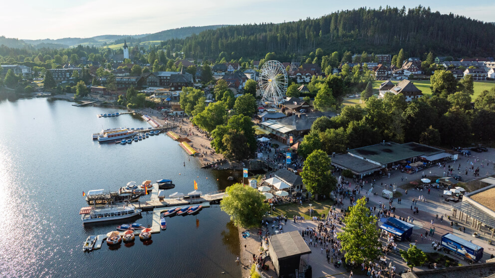 Luftaufnahme des Titisees mit Uferpromenade, Riesenrad und Sommerfest, umgeben von bewaldeten Hügeln.