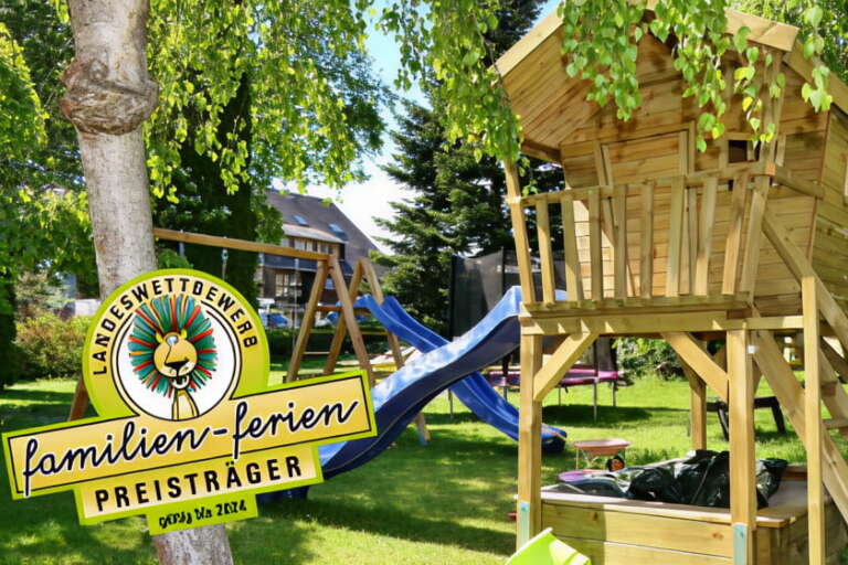 Ein hölzerner Spielplatz mit Baumhaus, Rutsche, Schaukel und Trampolin in einem grasbewachsenen Hof des Hotels Hochfirst, mit einer "familien-ferien"-Plakette im Vordergund.