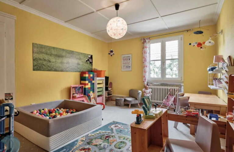 Kinderspielzimmer im H24 Hotel Hochfirst mit gelben Wänden, einem Bällebad, Spielzeug, Regalen, einer Spielküche, kleinen Stühlen, einem Schreibtisch und einem Fenster, durch das Tageslicht fällt.