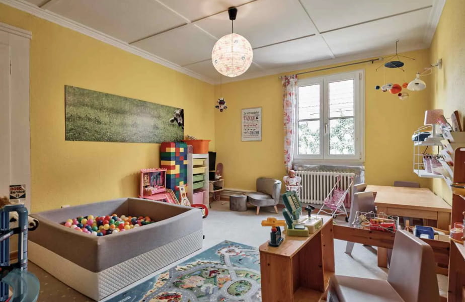 Kinderspielzimmer im H24 Hotel Hochfirst mit gelben Wänden, einem Bällebad, Spielzeug, Regalen, einer Spielküche, kleinen Stühlen, einem Schreibtisch und einem Fenster, durch das Tageslicht fällt.