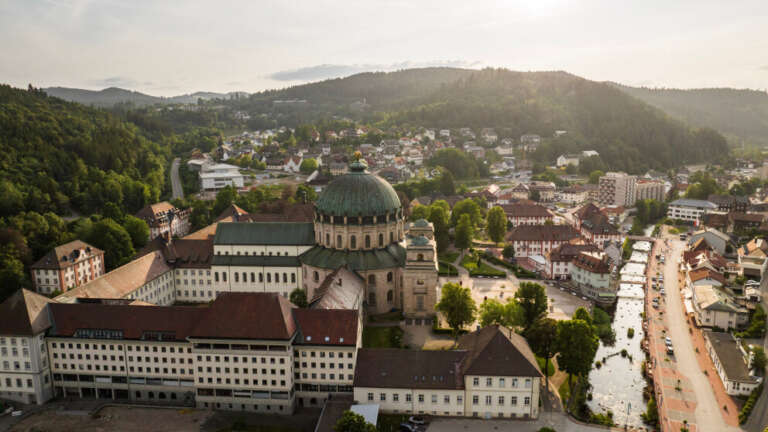 Luftaufnahme von St. Blasien im Schwarzwald mit markanter Kirchenkuppel im Zentrum, umgeben von Fachwerk- und Sandsteinhäusern, einem Fluss und bewaldeten Hügeln.