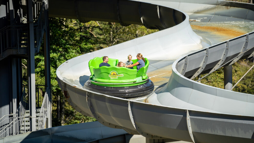 Familie sitzt in einem grünen Rundboot und fährt eine spiralförmige Wasserrutschbahn hinunter, umgeben von Bäumen.