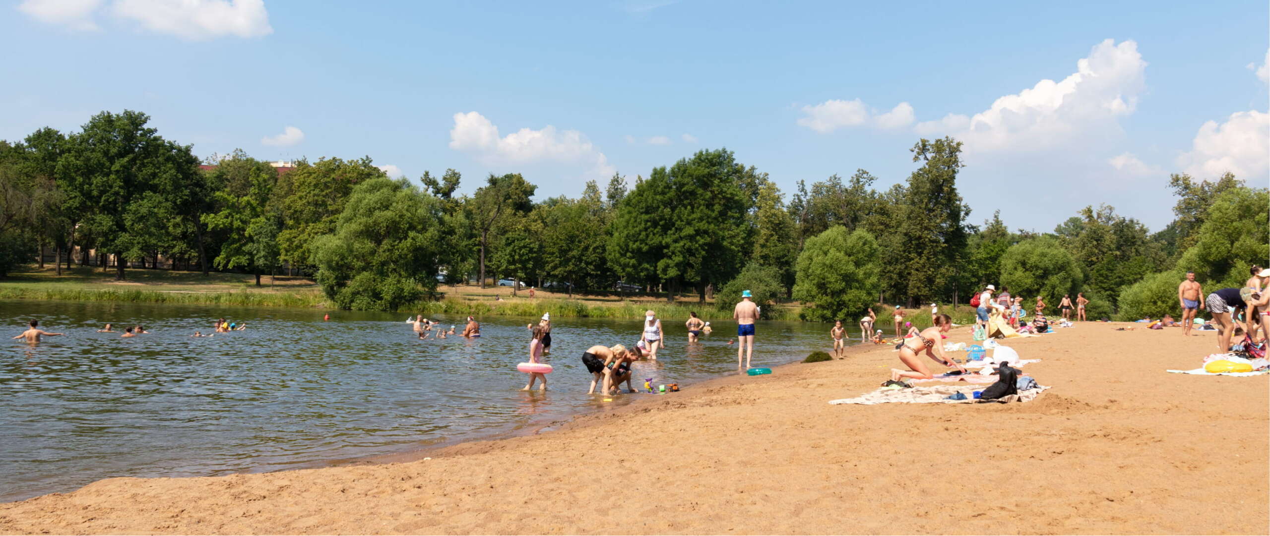 Sandstrand im Strandbad Grünau am Langen See: Badegäste im Sand und im flachen Wasser, Kinder mit Schwimmreifen am Ufer, dahinter bewaldetes Seeufer bei Sonnenschein.