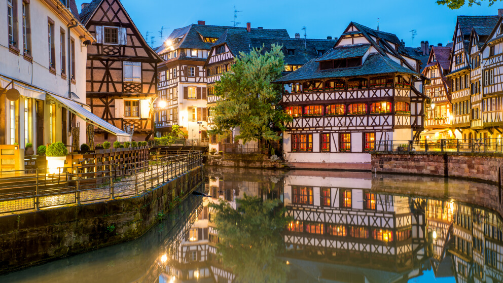 Beleuchtete Fachwerkhäuser am Kanal in Straßburg bei Dämmerung, spiegeln sich im ruhigen Wasser.