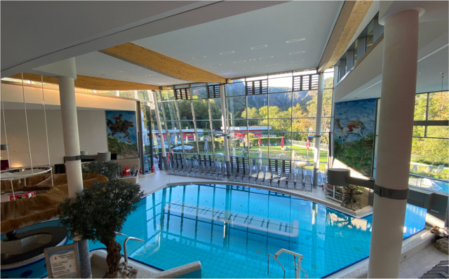 Großer Innenbereich der Therme mit einem Schwimmbecken, modernen Glasfronten mit Ausblick auf die grüne Berglandschaft und Wandmalereien mit Fantasie-Motiven.