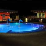 Beleuchteter Außenpool der Therme bei Nacht mit blauer Unterwasserbeleuchtung und Massagedüsen, umgeben von modernen Gebäuden mit roter Fassade und warmem Licht.