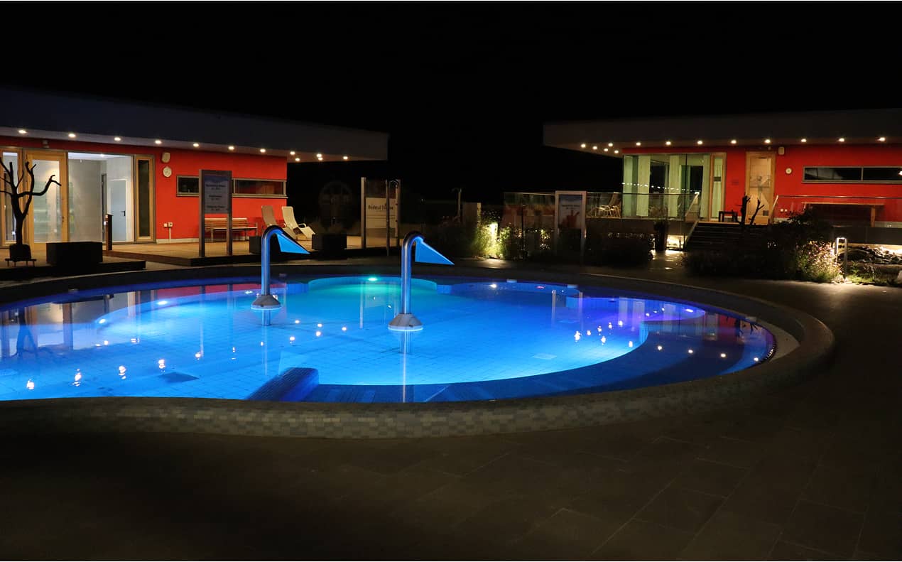 Beleuchteter Außenpool der Therme bei Nacht mit blauer Unterwasserbeleuchtung und Massagedüsen, umgeben von modernen Gebäuden mit roter Fassade und warmem Licht.