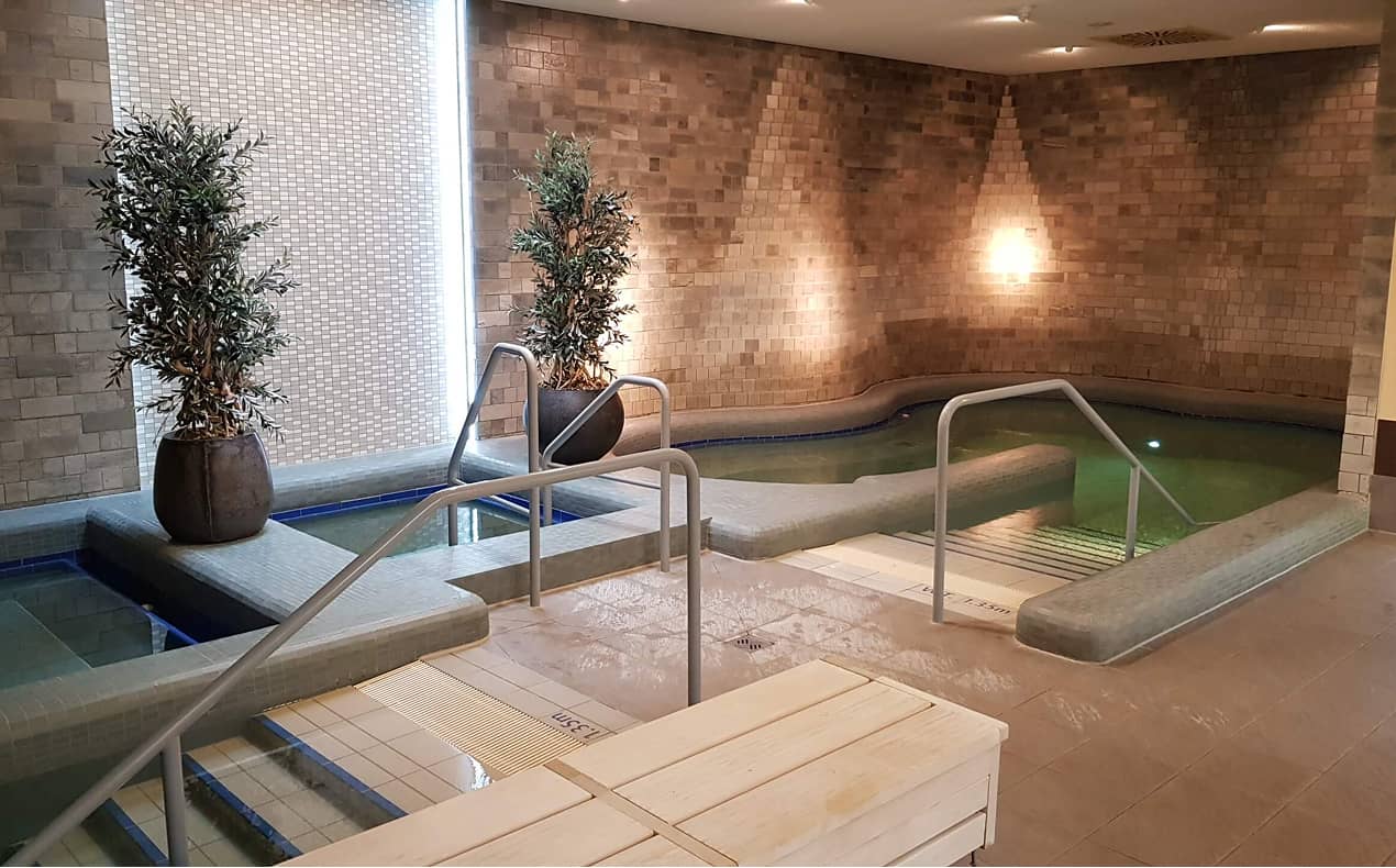 Innenbereich eines Spa-Pools mit warmen Licht, Pflanzen in großen Töpfen und einem Becken mit Handläufen zum Entspannen.