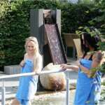 Zwei Frauen in blauen Handtüchern waten durch einen Kneipp-Pfad im Freien neben einem modernen Wasserbrunnen.