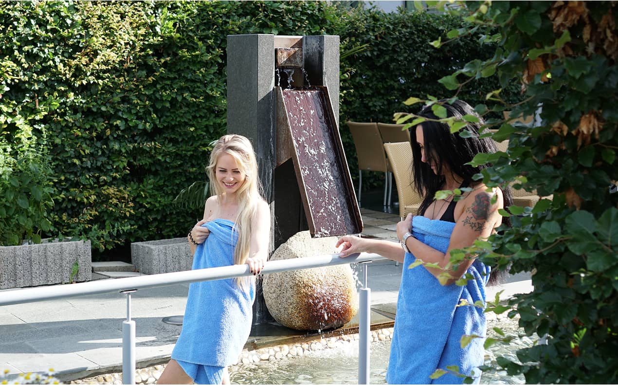 Zwei Frauen in blauen Handtüchern waten durch einen Kneipp-Pfad im Freien neben einem modernen Wasserbrunnen.