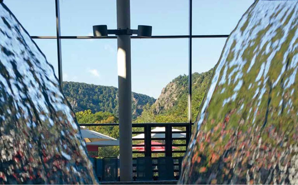 Blick durch zwei Wasserfälle im Innenbereich auf eine grüne Berglandschaft hinter großen Glasfenstern.
