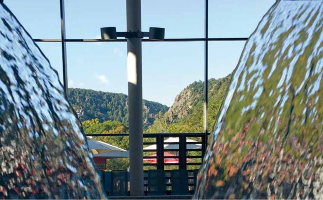 Blick durch zwei Wasserfälle im Innenbereich auf eine grüne Berglandschaft hinter großen Glasfenstern.