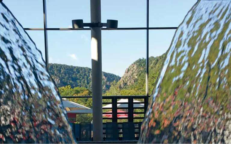 Blick durch zwei Wasserfälle im Innenbereich auf eine grüne Berglandschaft hinter großen Glasfenstern.