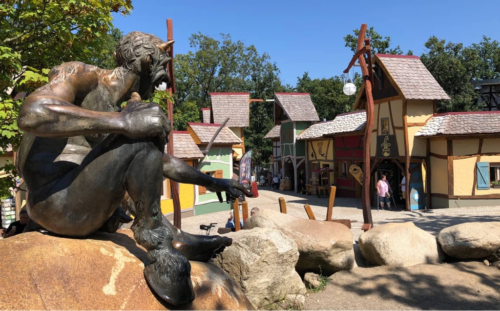 Bronze-Satyr-Statue sitzt auf Felsen, bunte Hexendorf-Kulisse im Hintergrund