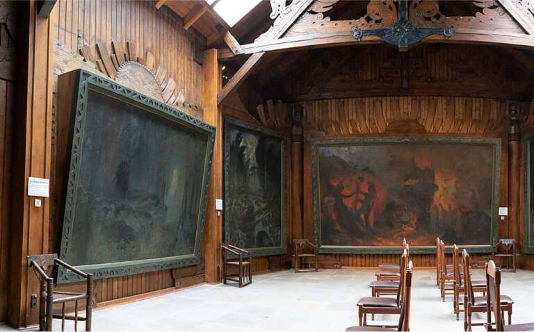 Großer, holzgetäfelter Saal mit riesigen düsteren Gemälden und Reihen von Holzstühlen