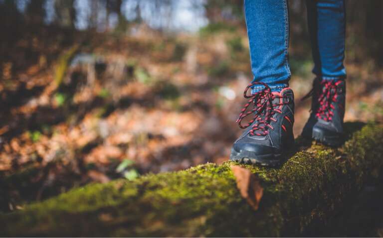 Wanderin mit blauen Jeans und schwarz-roten Wanderschuhen balanciert auf einem moosbewachsenen Baumstamm im herbstlichen Wald.