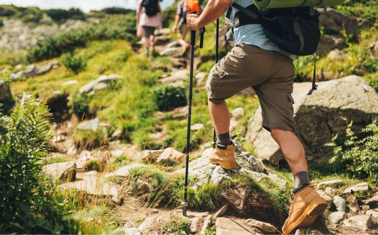 Gruppe von Wanderern in alpinem Gelände mit Wanderstöcken und Rucksäcken, ein Mann in Shorts und Wanderschuhen steigt über felsiges Gelände bergauf.