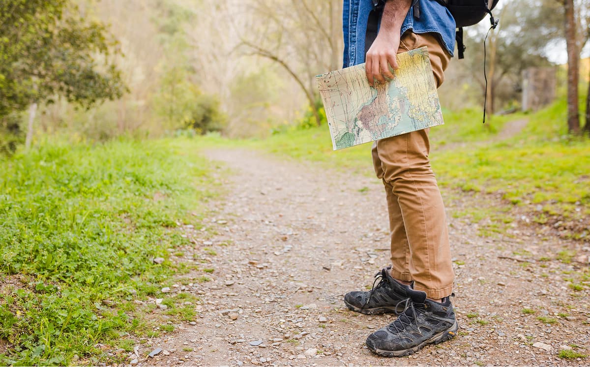 Wanderer mit Karte in der Hand steht auf einem breiten Kiesweg, umgeben von Wiese und Bäumen.