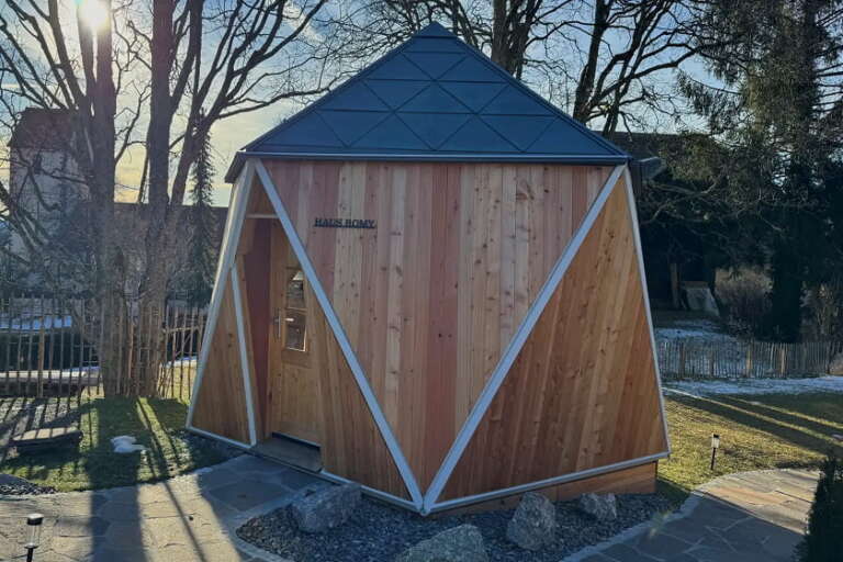 Eine kleine, moderne Holzhütte mit geometrischem Design und dreieckigem Dach steht in einem Garten, im Hintergrund sind Gras, Steine und Bäume zu sehen.