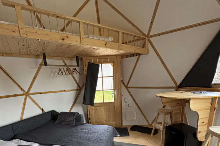 Innenansicht eines kleinen Tiny House in Form einer geodätischen Kuppel mit Holzakzenten, einem Bett, einem gemütlichen Hochbettbereich, einem runden Tisch mit Hockern, Kleiderhaken und einer Holztür, die nach draußen führt.