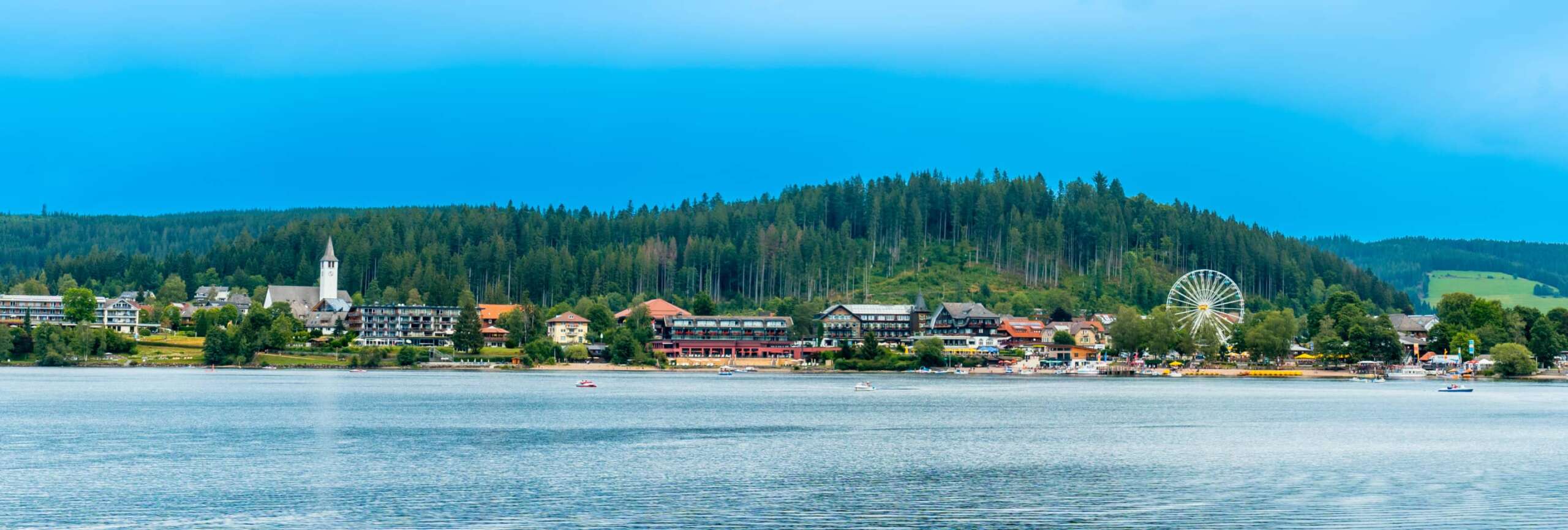 Panoramablick über den Titisee auf den Ort mit Kirche, Riesenrad und bewaldeten Hügeln im Schwarzwald