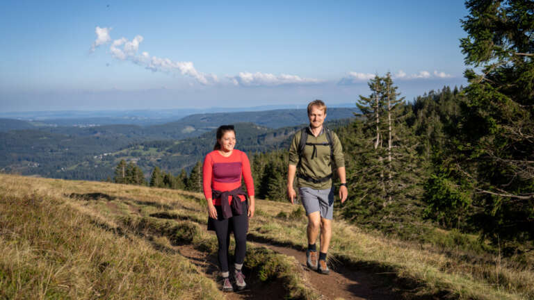 Frau und Mann wandern lächelnd auf einem Pfad über eine Bergwiese, im Hintergrund bewaldete Hügel und Fernsicht bis zum Horizont unter blauem Himmel mit Wolken.