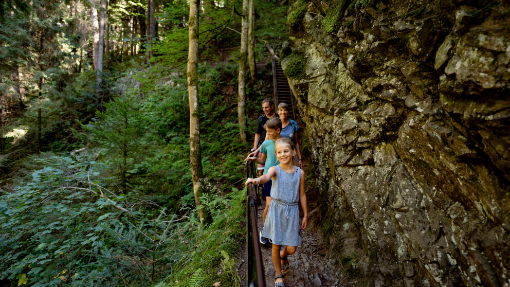 Familie mit zwei Kindern wandert an einem Geländer entlang eines schmalen Felspfads durch eine bewaldete Schlucht, im Hintergrund eine Treppe.