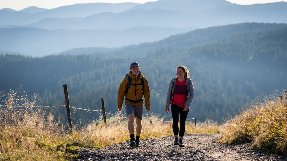 Mann und Frau wandern lächelnd auf einem Bergpfad, im Hintergrund bewaldete Hügelketten im Dunst der Morgensonne.
