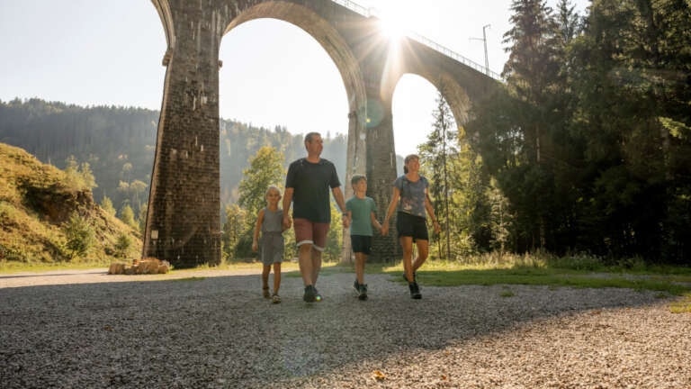 Familie mit zwei Kindern hält Händchen und geht auf einem Kiesweg unter einem hohen Eisenbahnviadukt, die Sonne scheint zwischen den Bögen hindurch.