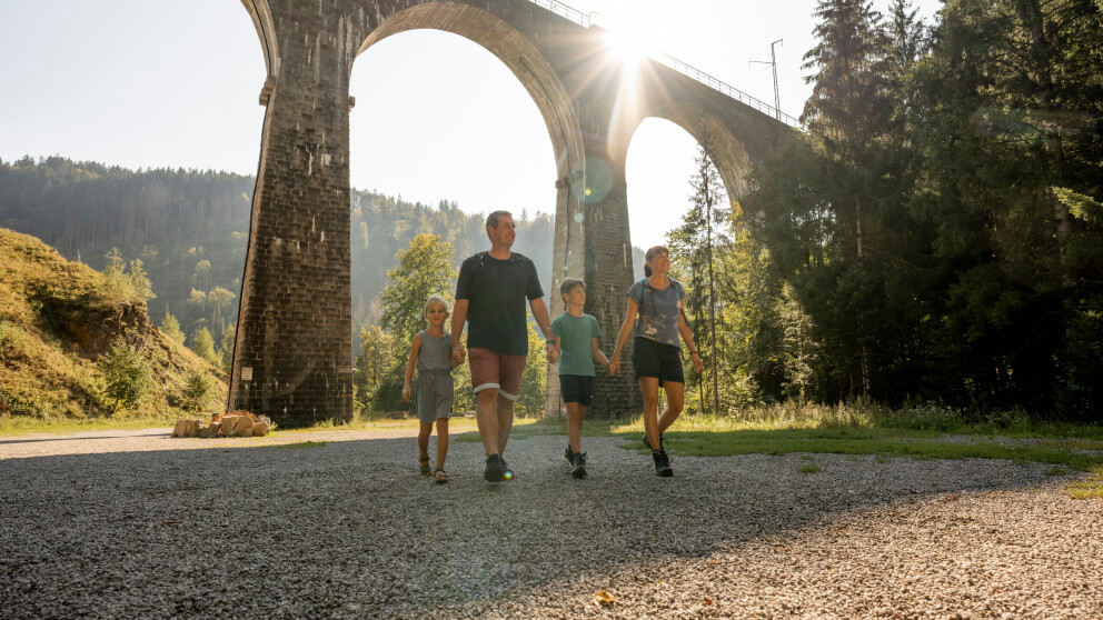 Familie mit zwei Kindern hält Händchen und geht auf einem Kiesweg unter einem hohen Eisenbahnviadukt, die Sonne scheint zwischen den Bögen hindurch.