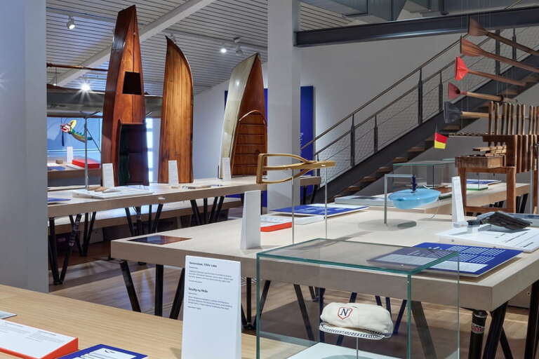 Ausstellungsraum mit Ausstellungs-Tischen, auf denen Modellboote, nautische Objekte und Informationskarten zu sehen sind; im Hintergrund sind Holzbootrümpfe und eine Treppe zu erkennen.